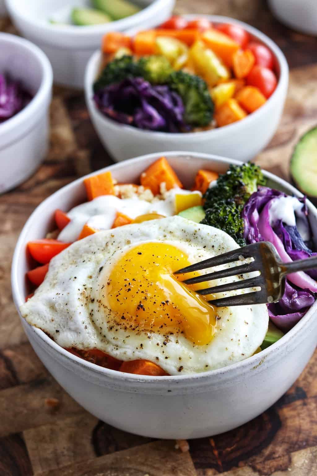 Up close shot of a fried egg being sliced with a fork and oozing onto the veggie bowl. 