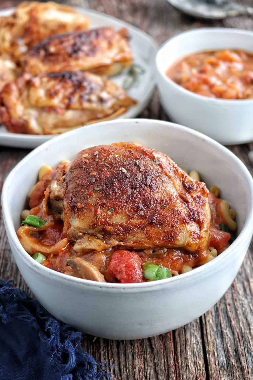 Chicken Cacciatore shown up close in a white bowl with spices and pasta with the extra chicken and sauce in bowls behind.