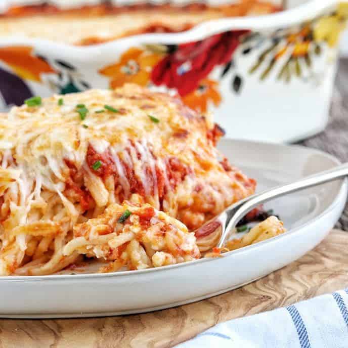 A slice of baked spaghetti on a white plate with a fork with the 9x13 pan with bright colored flowers on a white pan in the background.