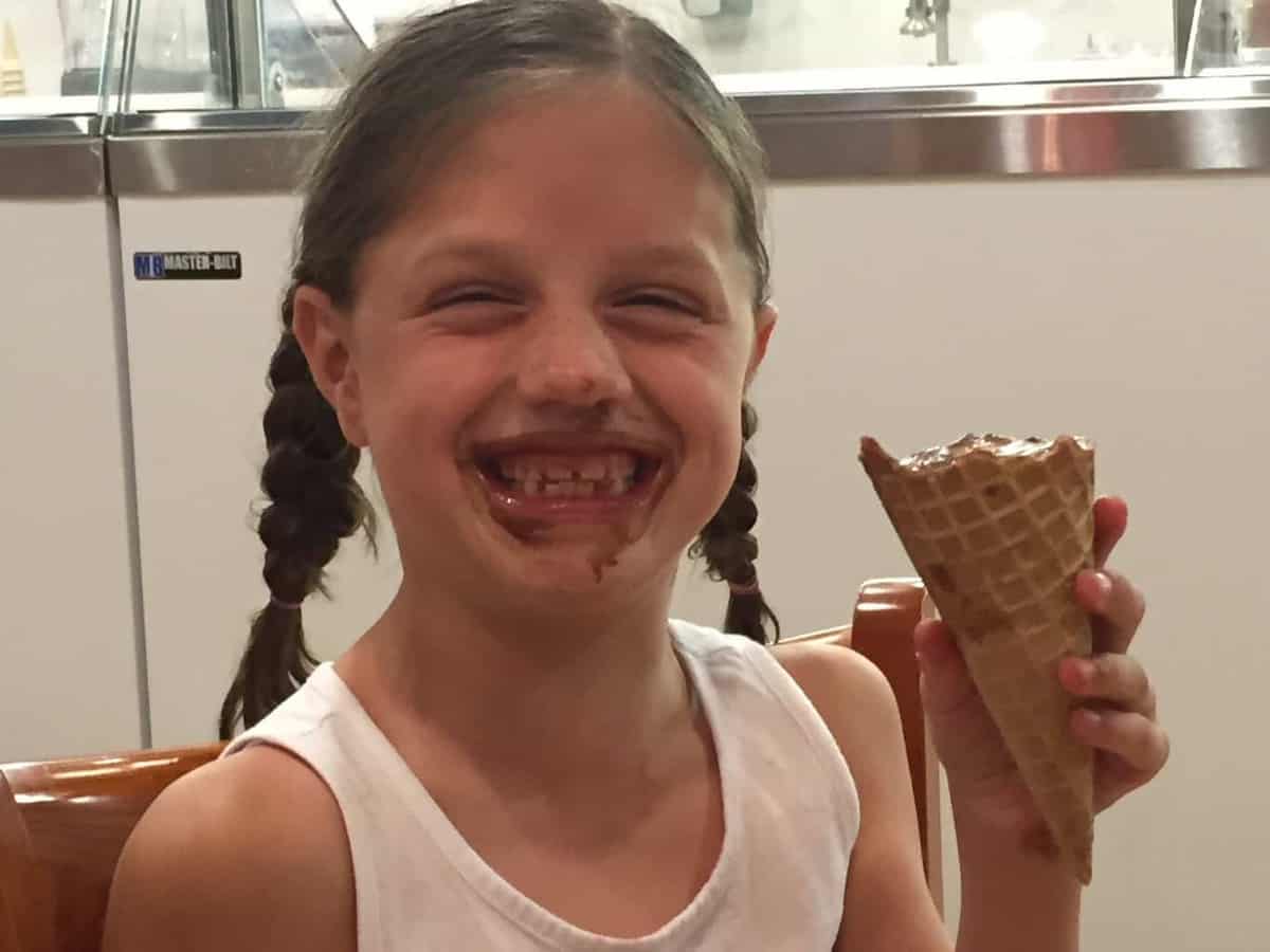 A young girl is smiling hugely covered in chocolate ice cream while holding the cone.