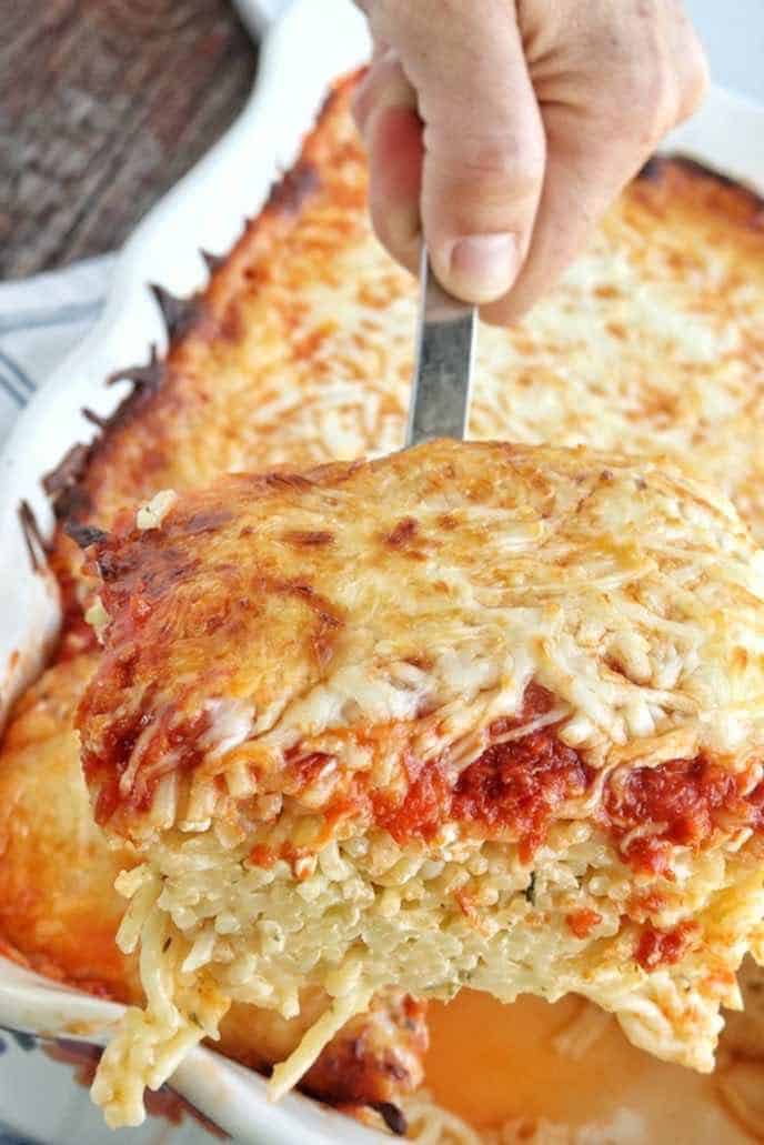 A pan of baked spaghetti with a square being lifted out of the pan by hand at an angle with a kitchen towel in the background.