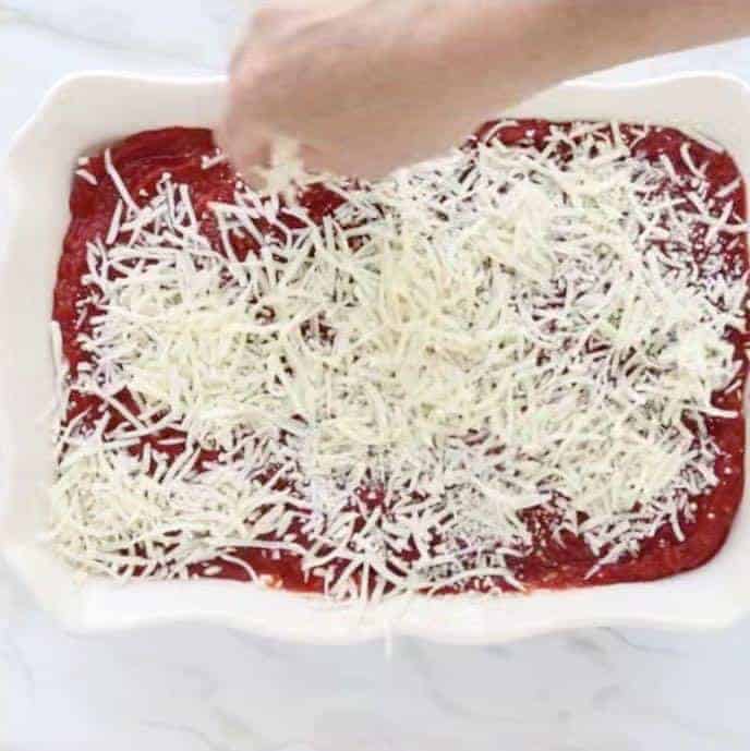Mozzarella cheese being sprinkled by hand over marinara sauce in a 9x13 inch white pan on a white marble table. 