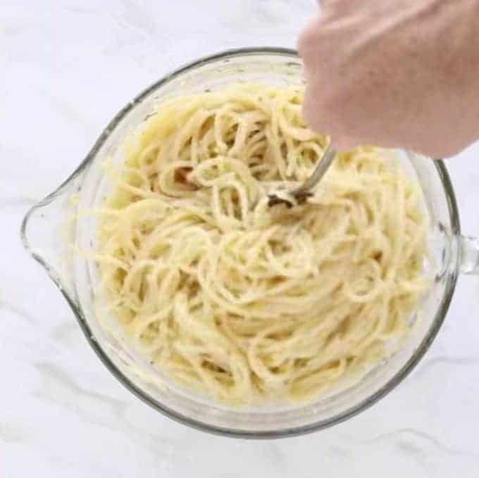 Pasta being stirred by hand in a clear glass bowl with a handle and spout on a marble table.
