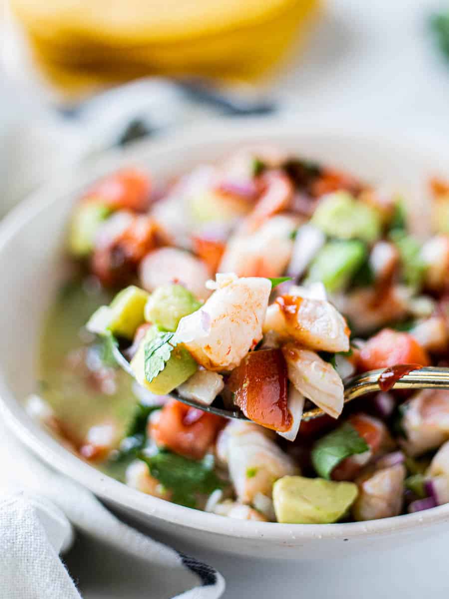 Diced shrimp, tomato, avocado, and cilantro on a gold spoon held close to the camera with the rest of the shrimp ceviche blurred behind.