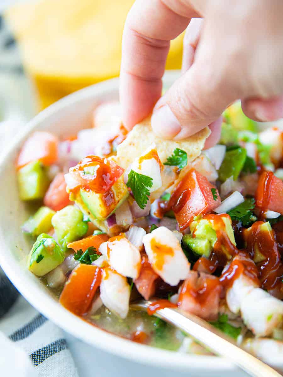 A hand is dipping a tortilla chip into a Mexican shrimp ceviche with avocado, tomatoes, and hot sauce.