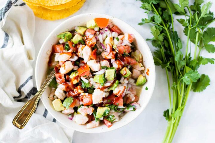 Shrimp Ceviche in a white bowl with a spoon with avocado, fresh tomatoes, cilantro, and hot sauce.
