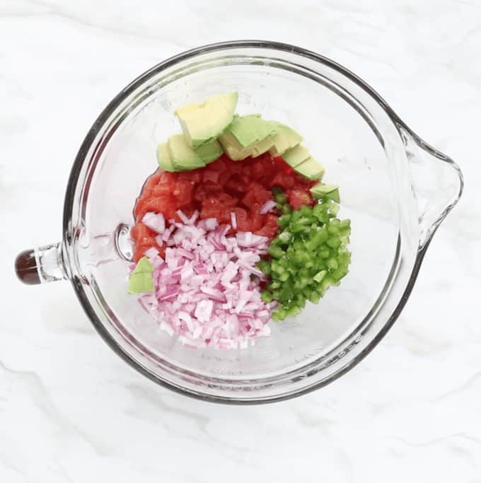 Canned tomatoes, diced avocado, diced red onions, and diced jalapeños in a clear medium sized bowl with a pour spout and handle