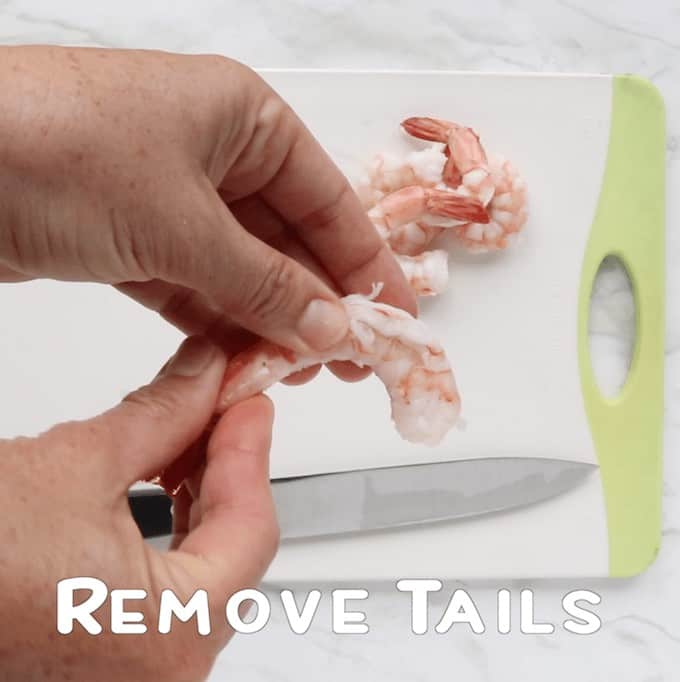 two hands removing tails from cooked shrimp with a cutting board and knife below