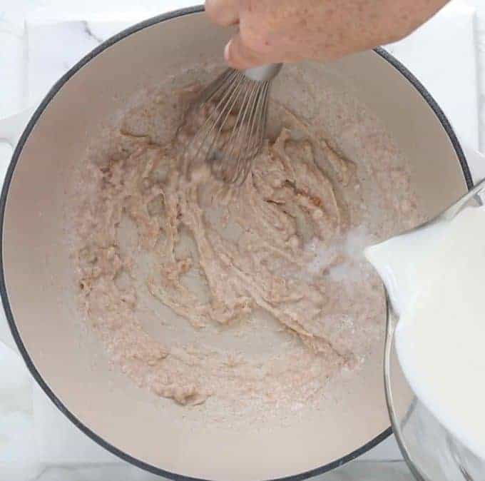 how to make Mac and cheese: top down view of white sauce pan with flour butter mixture thickened and milk being whisked in by hand.