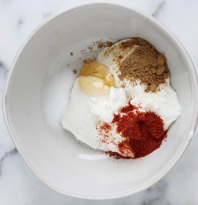 A white bowl with yogurt, chili powder, cumin, honey and salt on a white marble surface