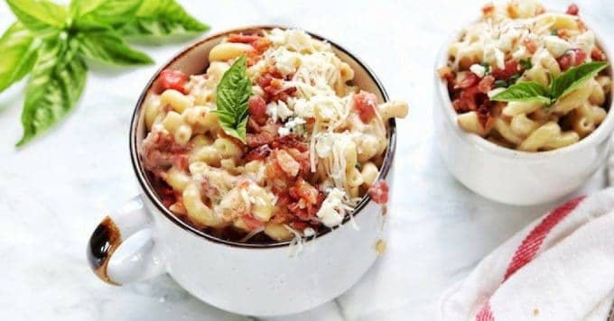 Mac and cheese with bacon in a white bowl with a handle topped with bacon bites and more cheese.