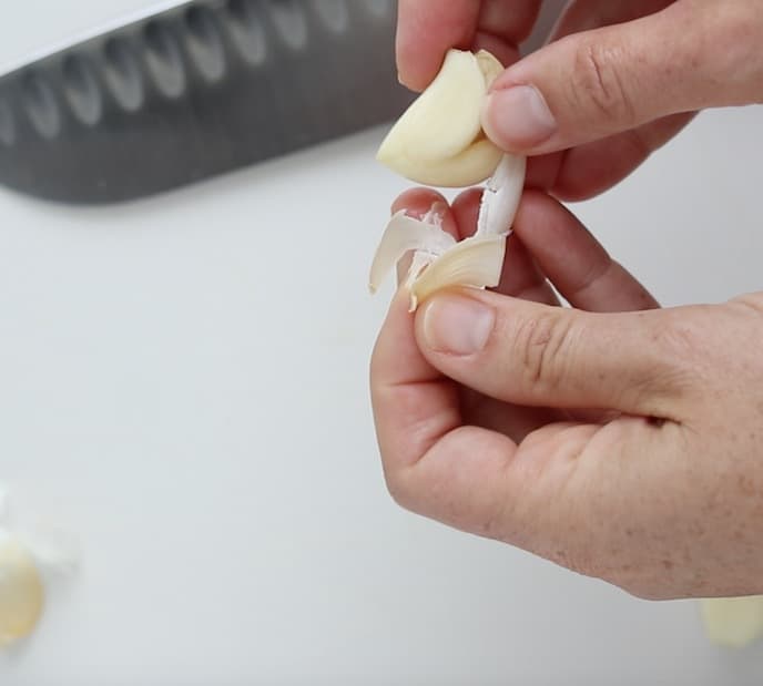 A clove of garlic being separated from the skin by two hands with a chefs knife in the background on a white cutting board