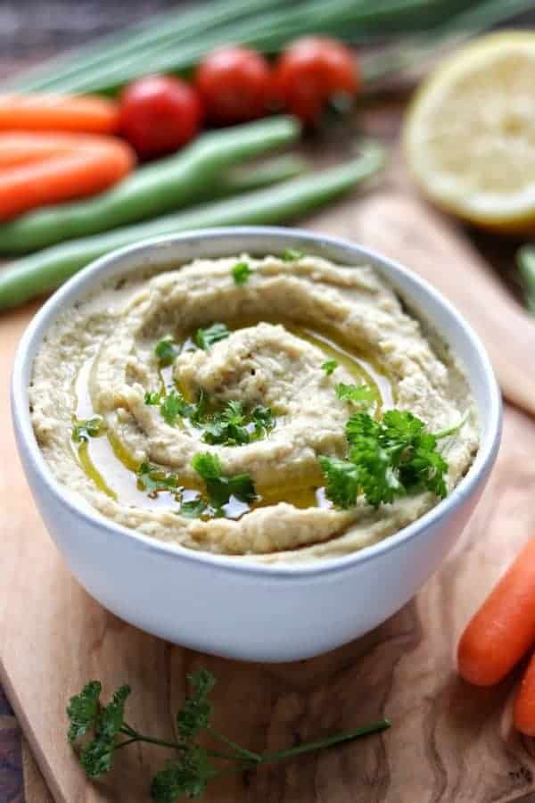 A white bowl filled with ranch hummus on top of a wood plank with various vegetables around the bowl. 