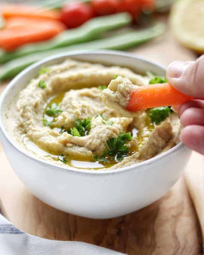 A white bowl filled with hummus shown on top of a wood plank with a hand dipping a carrot into the bowl of hummus. 