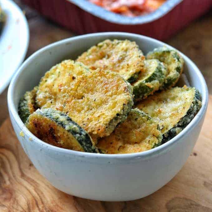 Zucchini crisps are shown stacked in a white bowl on a wooded surface.