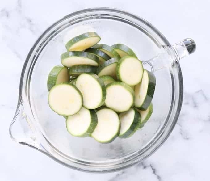  raw sliced zucchini is shown in a clear white bowl on a white marble surface. 