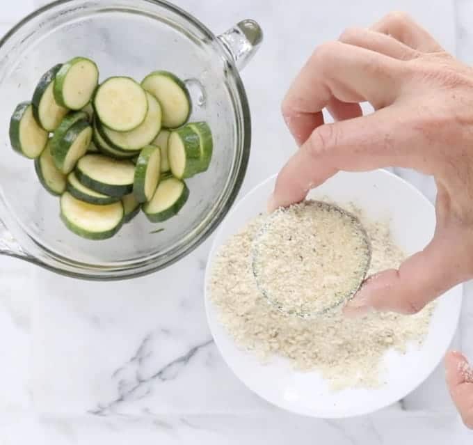 raw zucchini round is coated in panko crumbs next to a bowl of zucchini slices and panko crumbs on a white marble surface.