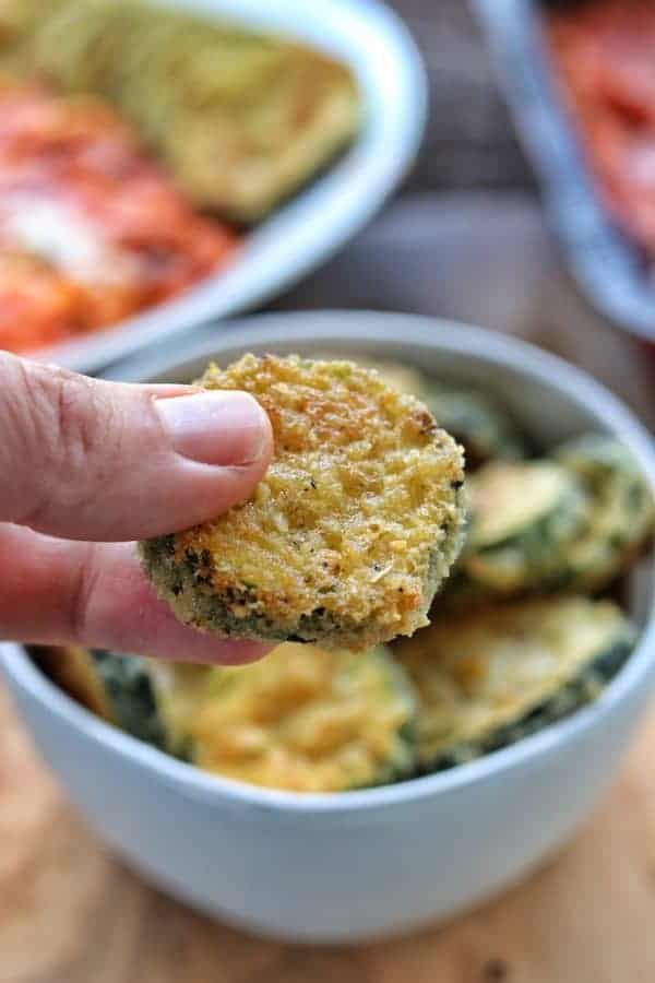 baked zucchini squash coated is held by a hand up close with the bowl of zucchini in the background.