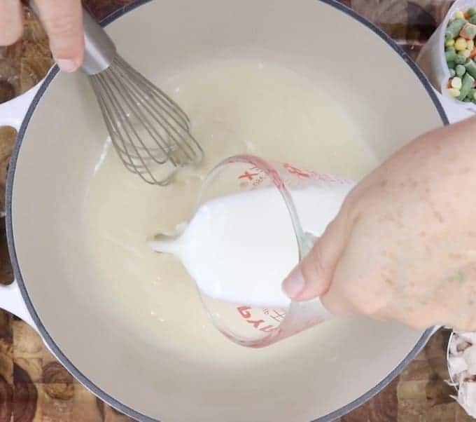 Milk is being whisked into the flour butter and broth mixture in a white pot for easy chicken pot pie with pie crust.