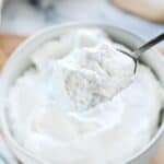 Stablilized whipped cream shown in a white bowl with a spoon scooping some out.