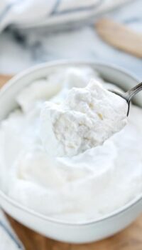 Stablilized whipped cream shown in a white bowl with a spoon scooping some out.