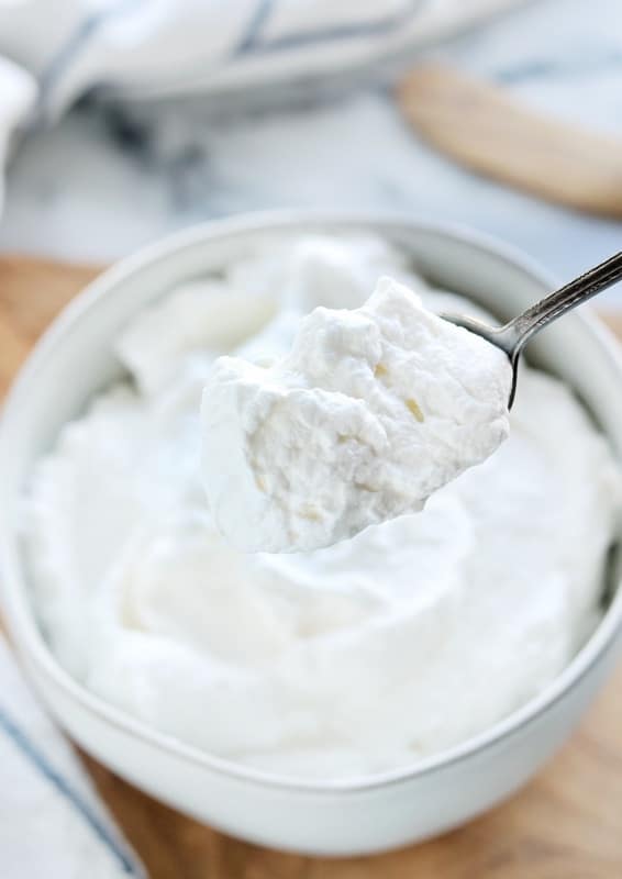  Recette de crème fouettée stabilisée présentée sur une cuillère tenue au-dessus d'un bol blanc rempli de crème fouettée maison sur une surface blanche.