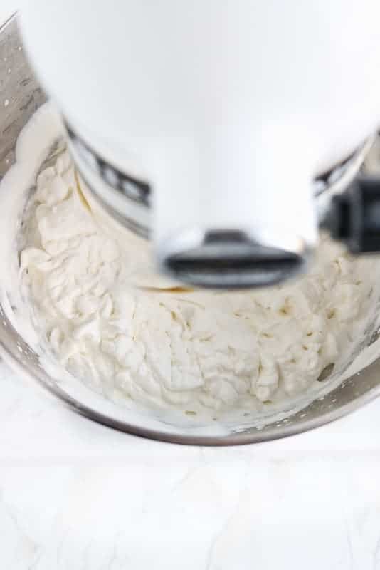 Crème fouettée stabilisée montrée de haut en bas d'un mélangeur d'aide à la cuisine blanc montrant de la crème fouettée à pointe douce à moyenne dans un bol à mélanger en métal.