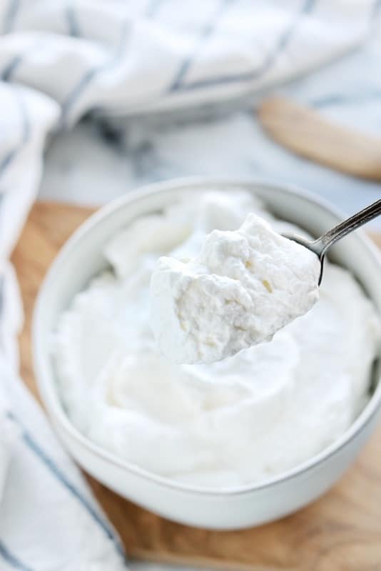 Crème fouettée stabilisée montrée sur une cuillère de près avec un bol blanc avec de la crème fouettée stabilisée en arrière-plan.