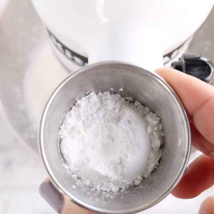 Crème fouettée stabilisée en cours de fabrication, montrant du sucre en poudre dans un petit bol en métal devant un mélangeur blanc.