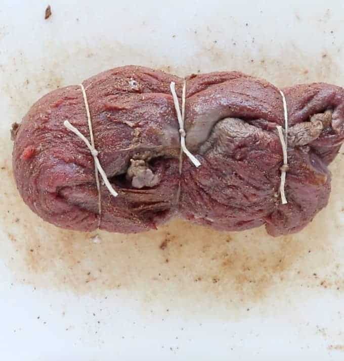 whole beef tenderloin wrapped into itself and tied three times with cooking tine all on a white cutting board surface.