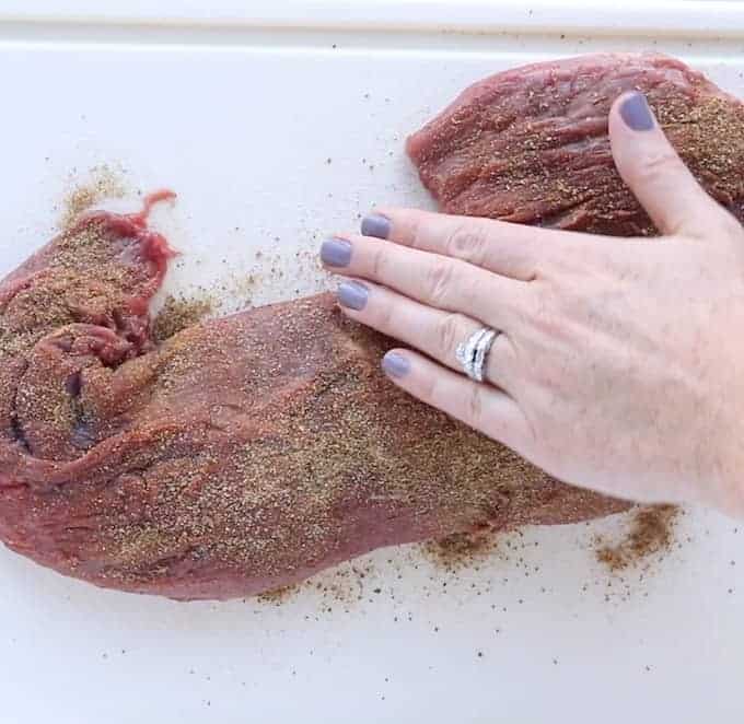 raw whole beef tenderloin is being seasoned with a hand rubbing the spices into the meat on a white cutting board.