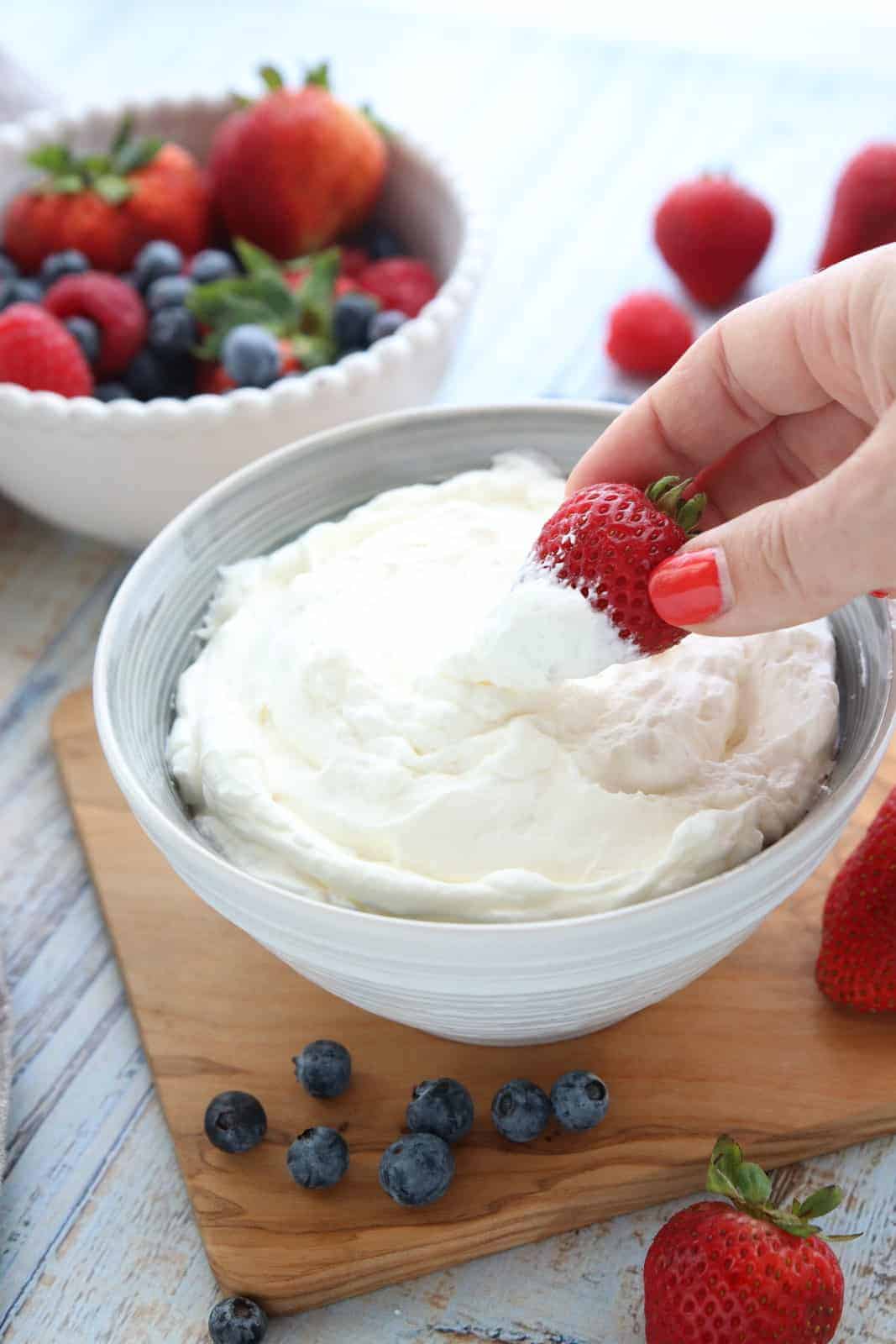 Long lasting whipped cream shown with berries being dipped in the bowl of whipped cream that's sitting on a wooden surface.