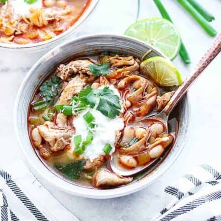 A bowl of chicken chili shown in a bowl with white beans, chicken celery and some sour cream and fresh green onions on top.