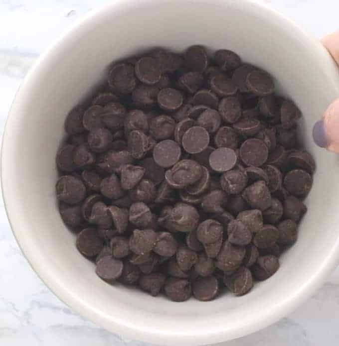 Chocolate chips in a white glass bowl on a white surface.