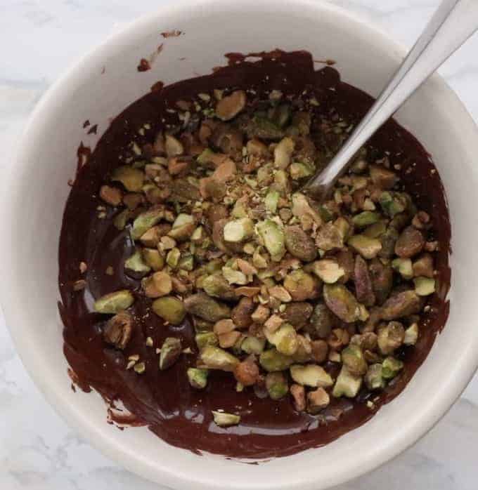 Melted chocolate in a white glass bowl with a spoon stirring in crushed pistachios.