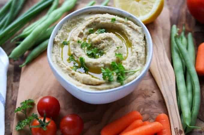 A white bowl filled with ranch hummus on top of a wood plank with various vegetables surrounding the bowl. 