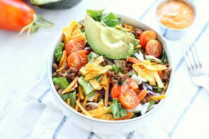 Healthy taco salad recipe shown in a white bowl with fresh tomatoes and avocados on top with tortilla crisps all on a white surface.