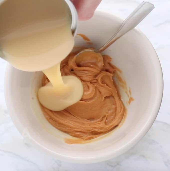 Easy Peanut butter fudge being made in a white bowl with peanut butter and a spoon in it with sweetened condensed milk being poured in. 