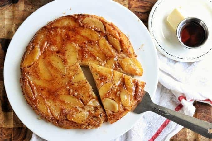 An apple cinnamon pancake being shown on a white plate with a spatula serving a slice. A dish of butter and syrup are next to the pancake all on top of a wooden countertop.