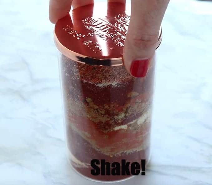 Chicken taco seasoning mix recipe shown in a clear glass jar with lid before being mixed by shaking on a white surface. 