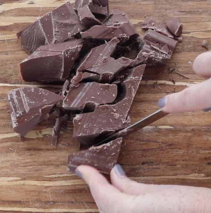 A knife breaking apart a large chunk of milk chocolate on a wooden cutting board. 