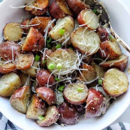 A white bowl with roasted red potatoes sliced in half with shredded parmesan and green onion on top.
