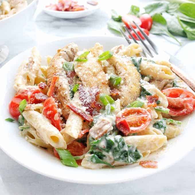 A white bowl with creamy chicken pasta with bacon, tomatoes, and spinach.