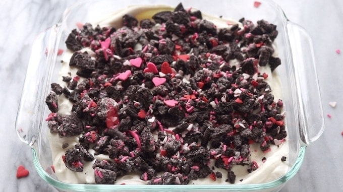 Oreo Dirt Cake being assembled showing crushed Oreos in a 9x9 clear glass pan with pudding mix and more Oreos on top.