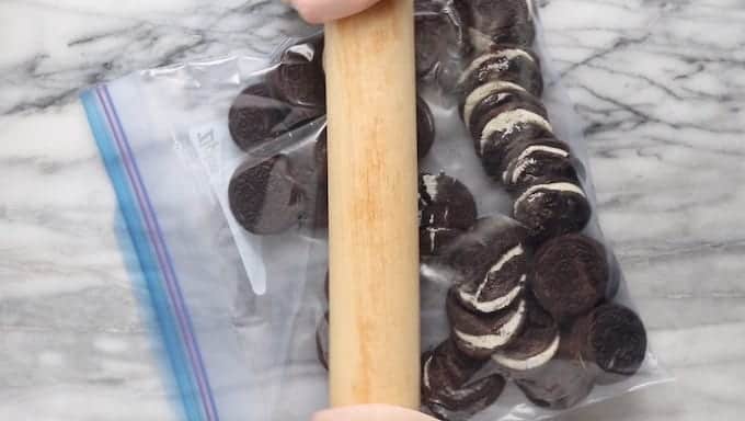 Oreo dirt cake being made by showing Oreos in ziplock bag on white marble surface with a rolling pin crushing them.