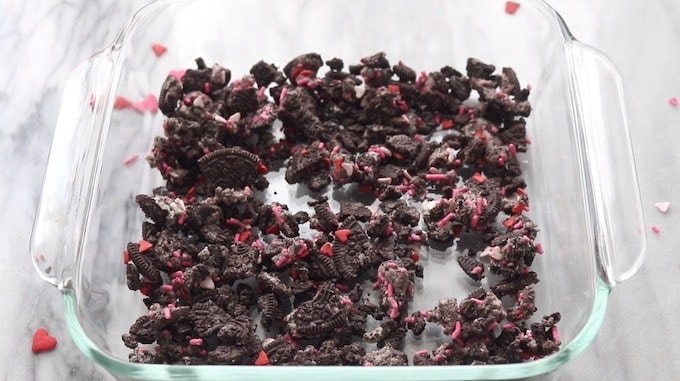 Oreo Dirt Cake being assembled showing crushed Oreos in a 9x9 clear glass pan on a white marble surface.