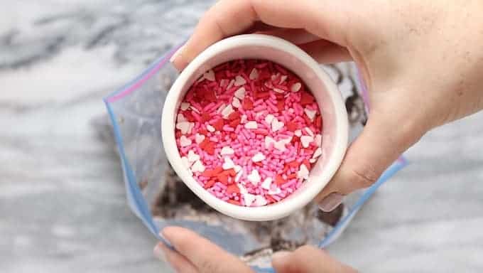 Oreo dirt cake being made showing valentines day sprinkles being added to a ziplock bag of crushed Oreo cookies in a ziplock bag.