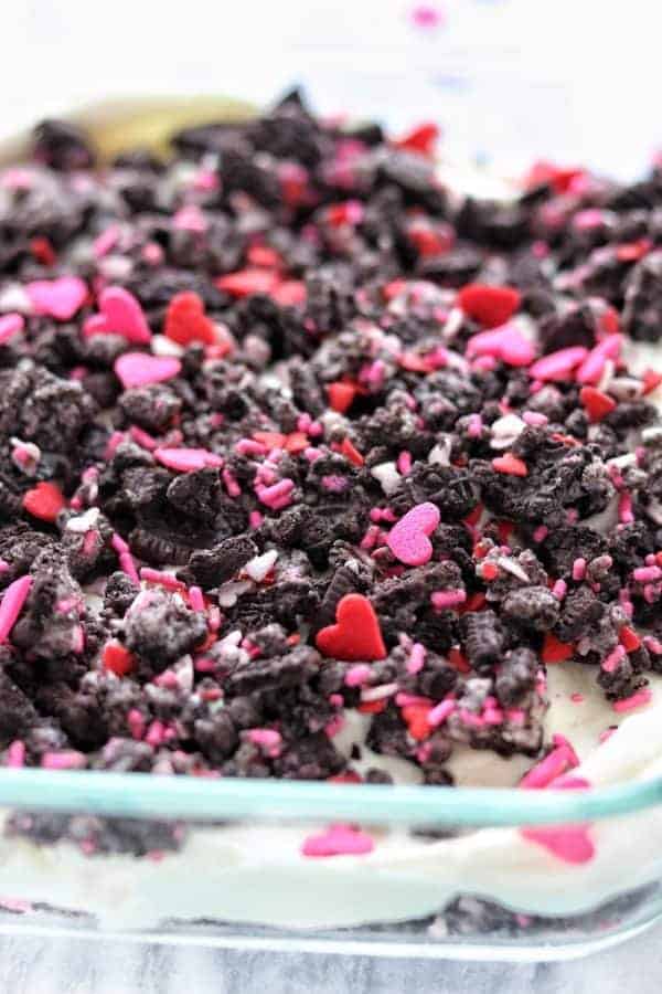 Dirt pudding recipe shown in a clear glass pan topped with crushed Oreos and pink and red heart sprinkles on a white surface.