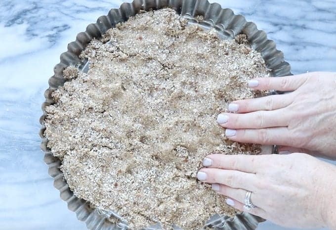Fruit flan, a healthy dessert idea being made, showing the oat/date mixture being spread into a metal circle flan pan with 2 hands forming a crust.