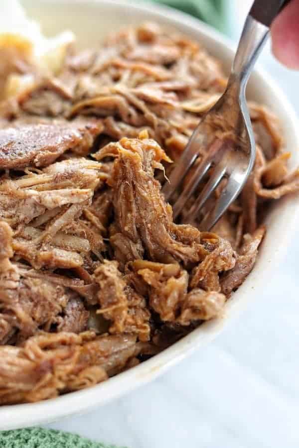 pork shoulder roast in pressure cooker shown close up in a white bowl with a fork diving into the instant pot pork on white marble surface. 